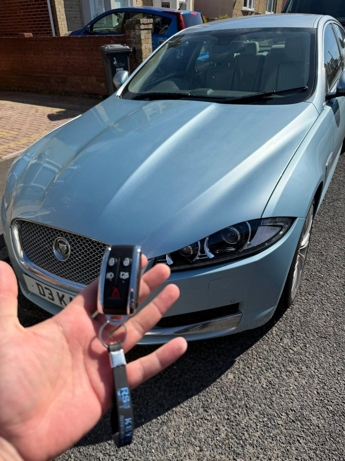 Person holding a car key fob in front of a silver Jaguar sedan parked on a driveway