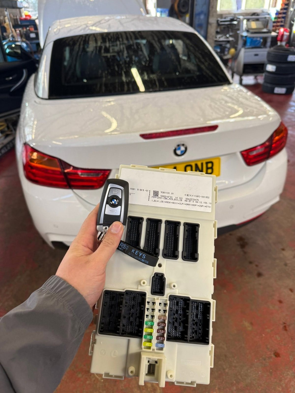 Person holding BMW car key fob and fuse box in front of white BMW vehicle in auto repair shop