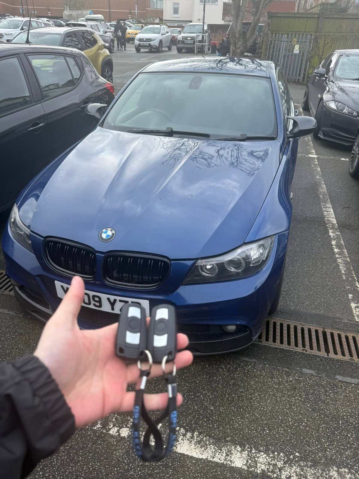 Person holding car keys in front of a blue BMW parked in a lot with other vehicles